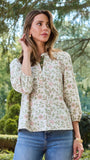 A woman wearing a long-sleeve button-up blouse with a delicate pink and green floral pattern on a cream background, paired with blue jeans, standing outdoors in front of a white column and green trees.