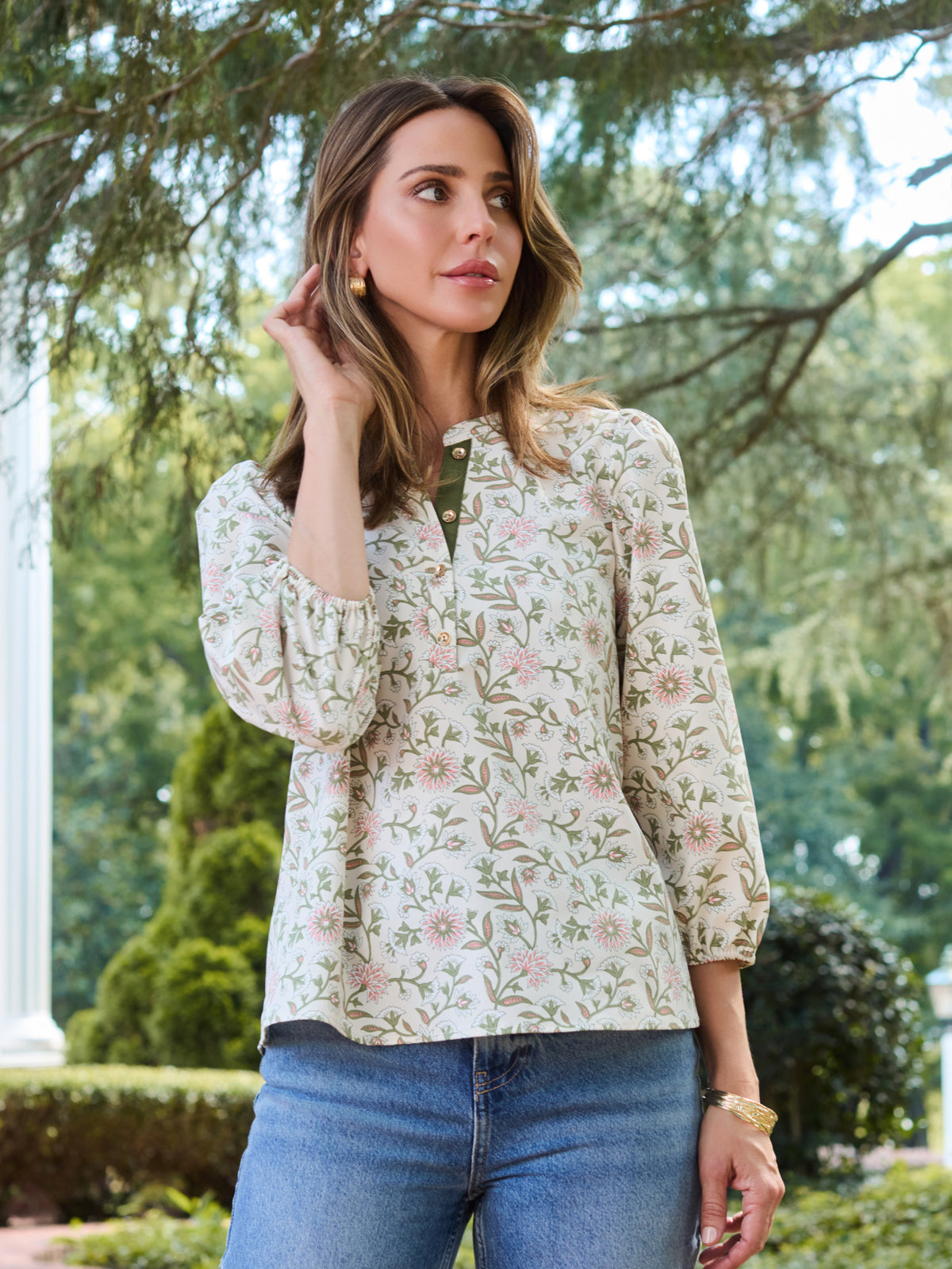 A woman wearing a long-sleeve button-up blouse with a delicate pink and green floral pattern on a cream background, paired with blue jeans, standing outdoors in front of a white column and green trees.