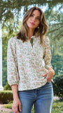 A woman wearing a long-sleeve button-up blouse with a delicate pink and green floral pattern on a cream background, paired with blue jeans, standing outdoors in front of a white column and green trees.