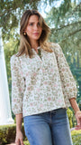 A woman wearing a long-sleeve button-up blouse with a delicate pink and green floral pattern on a cream background, paired with blue jeans, standing outdoors in front of a white column and green trees.