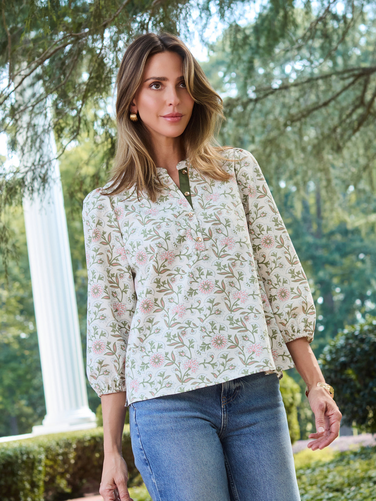 A woman wearing a long-sleeve button-up blouse with a delicate pink and green floral pattern on a cream background, paired with blue jeans, standing outdoors in front of a white column and green trees.