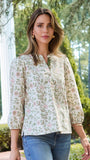 A woman wearing a long-sleeve button-up blouse with a delicate pink and green floral pattern on a cream background, paired with blue jeans, standing outdoors in front of a white column and green trees.
