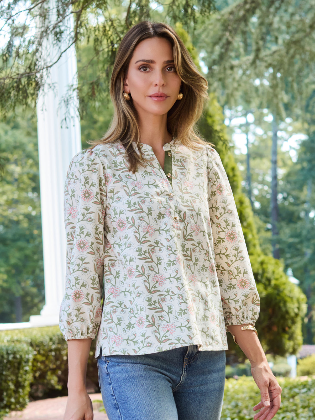 A woman wearing a long-sleeve button-up blouse with a delicate pink and green floral pattern on a cream background, paired with blue jeans, standing outdoors in front of a white column and green trees.