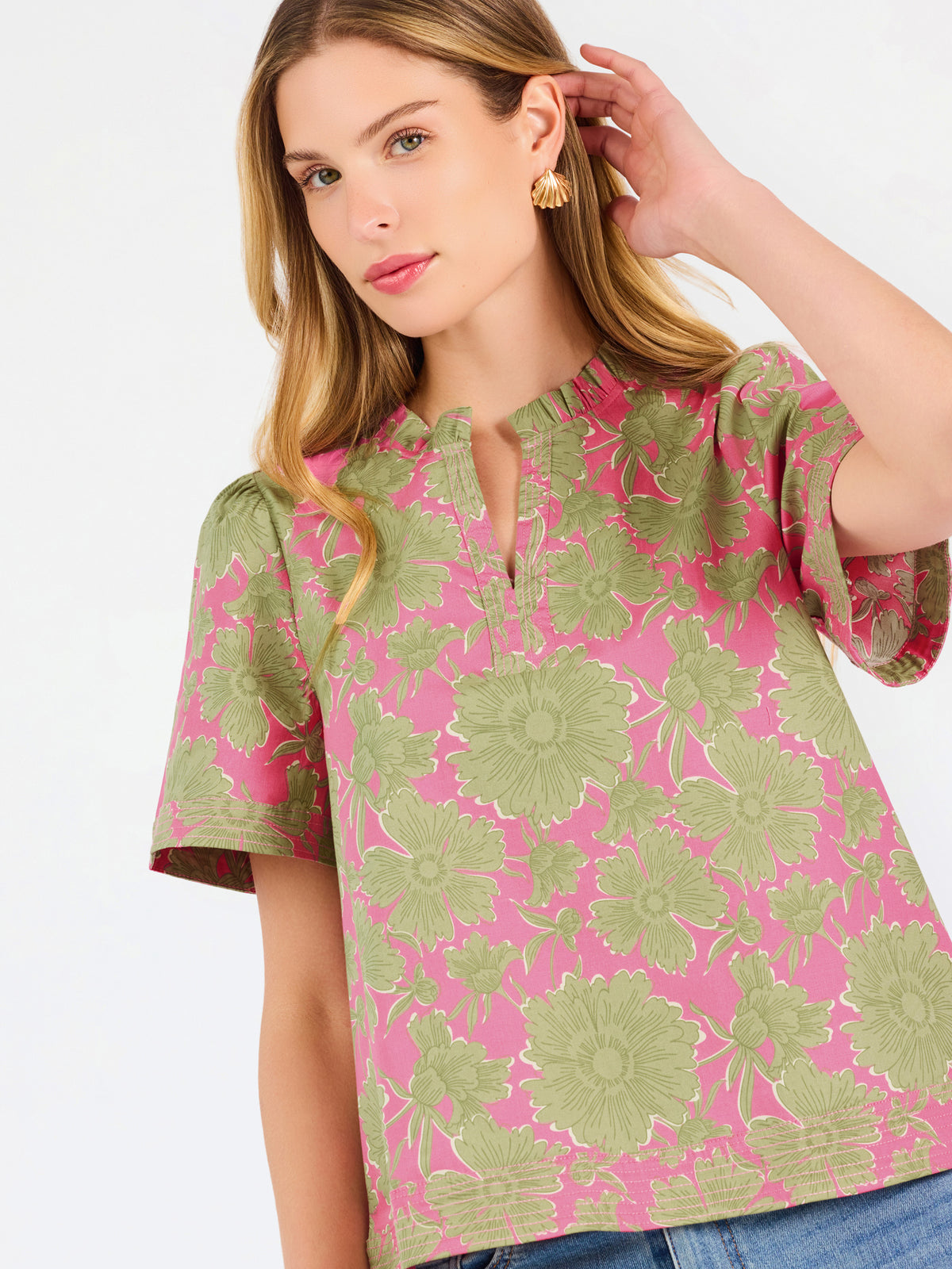 A woman modeling a short-sleeve top with a pink and olive green floral pattern, paired with blue jeans, against a plain white studio background.