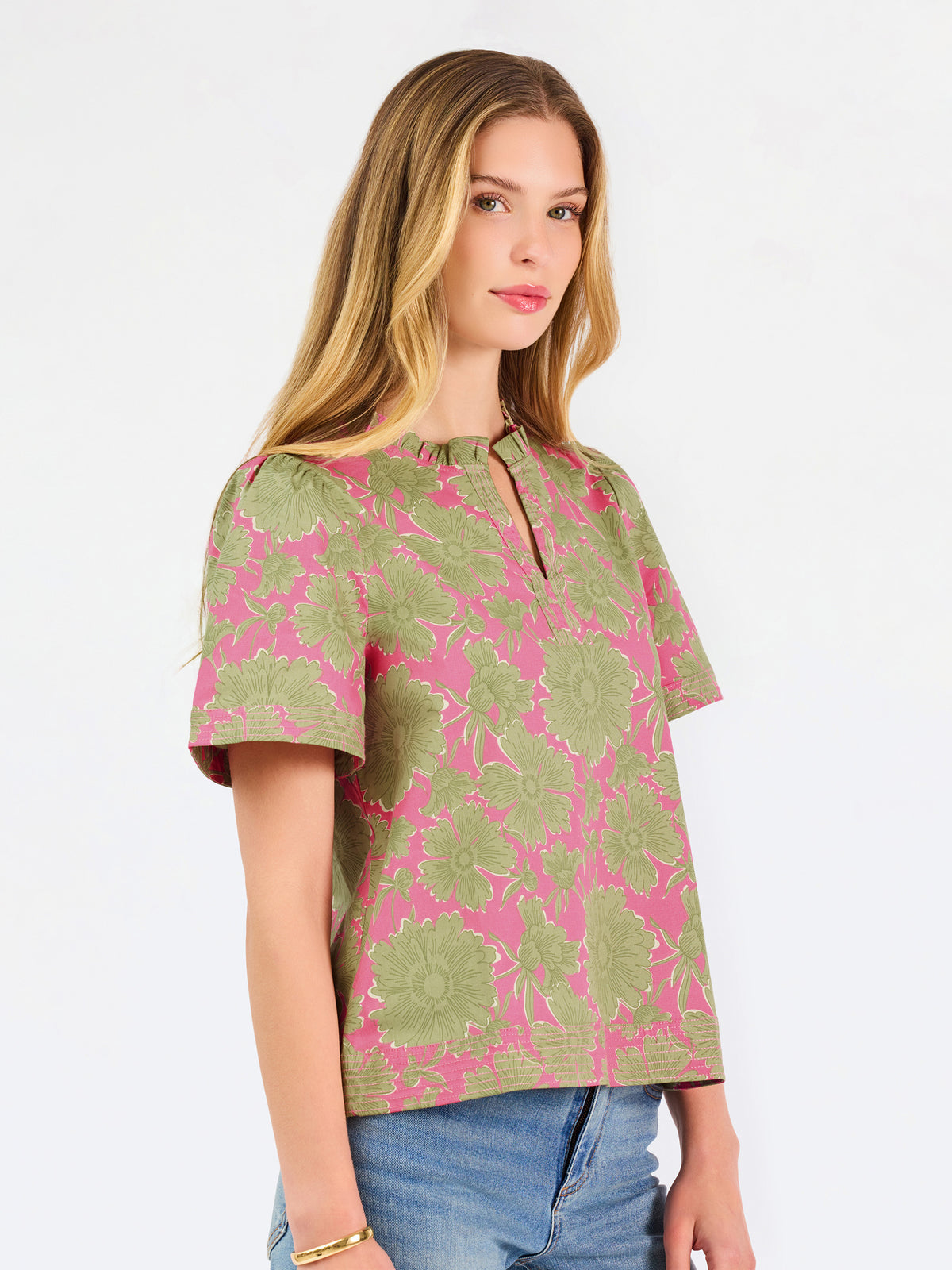 A woman modeling a short-sleeve top with a pink and olive green floral pattern, paired with blue jeans, against a plain white studio background.