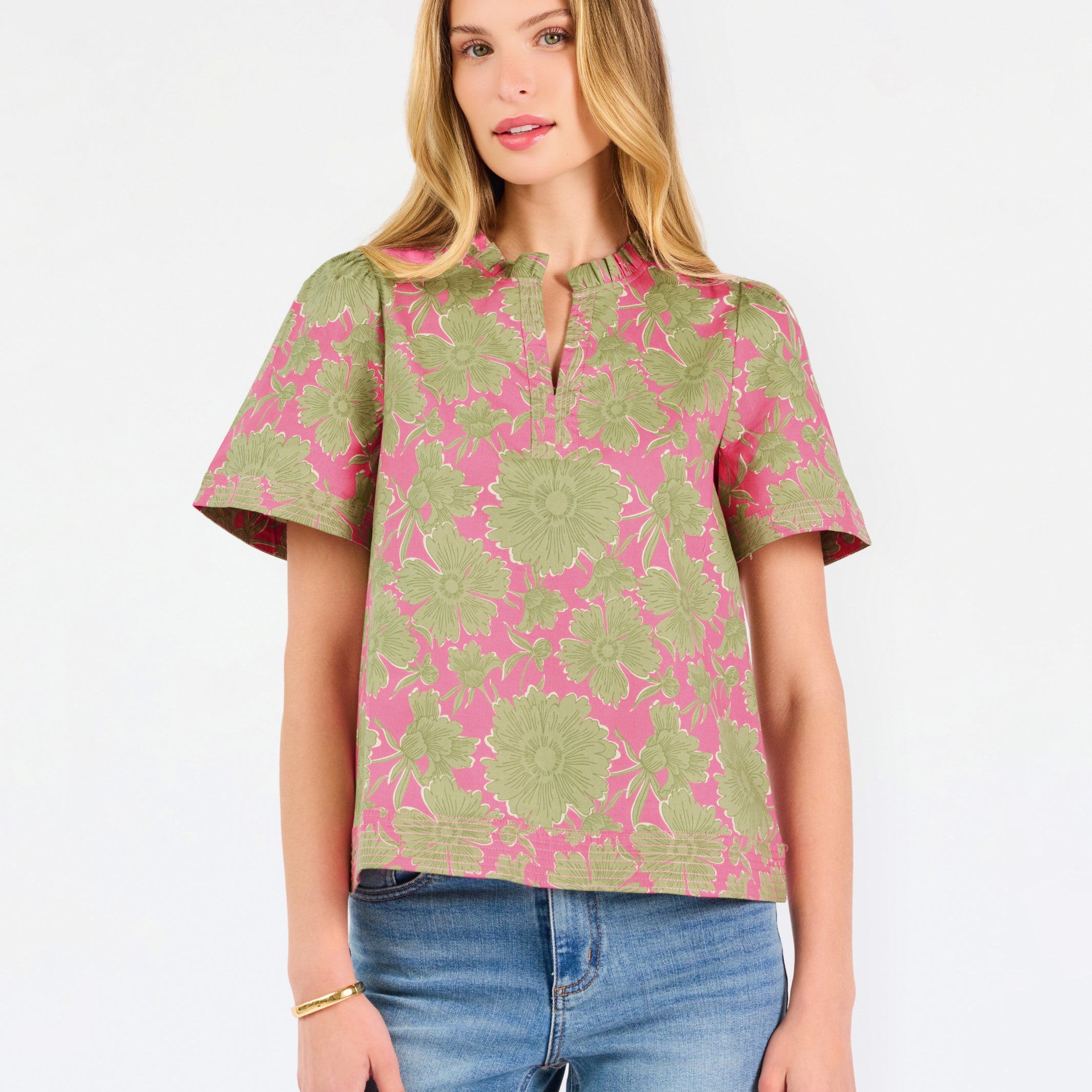 A woman modeling a short-sleeve top with a pink and olive green floral pattern, paired with blue jeans, against a plain white studio background.