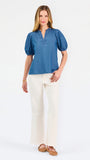 A woman modeling a blue short-sleeve puff-sleeve blouse with a split neckline, paired with cream wide-leg pants and brown flat sandals, against a plain white studio background.