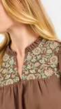 A woman modeling a brown short-sleeve blouse with a floral-patterned yoke and split neckline, paired with blue jeans, against a plain white background.