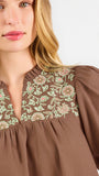 A woman modeling a brown short-sleeve blouse with a floral-patterned yoke and split neckline, paired with blue jeans, against a plain white background.