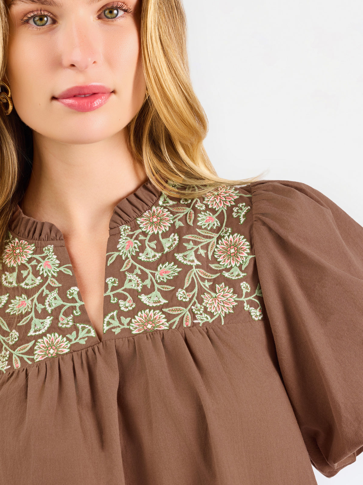 A woman modeling a brown short-sleeve blouse with a floral-patterned yoke and split neckline, paired with blue jeans, against a plain white background.