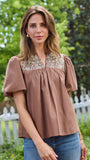 A woman wearing a brown short-sleeve blouse with a floral embroidered yoke and blue jeans, standing outdoors in front of a white picket fence and green trees.