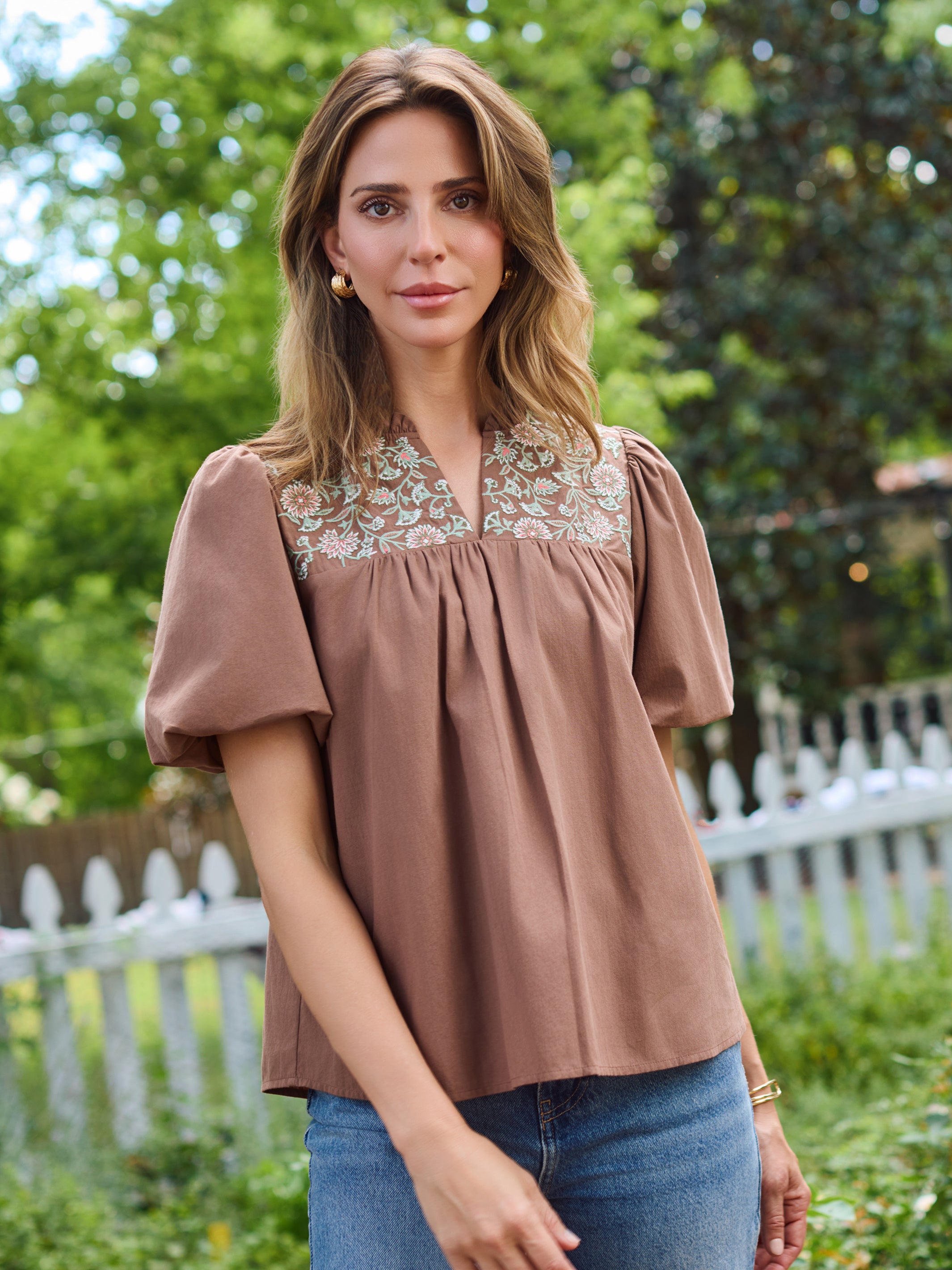 A woman wearing a brown short-sleeve blouse with a floral embroidered yoke and blue jeans, standing outdoors in front of a white picket fence and green trees.