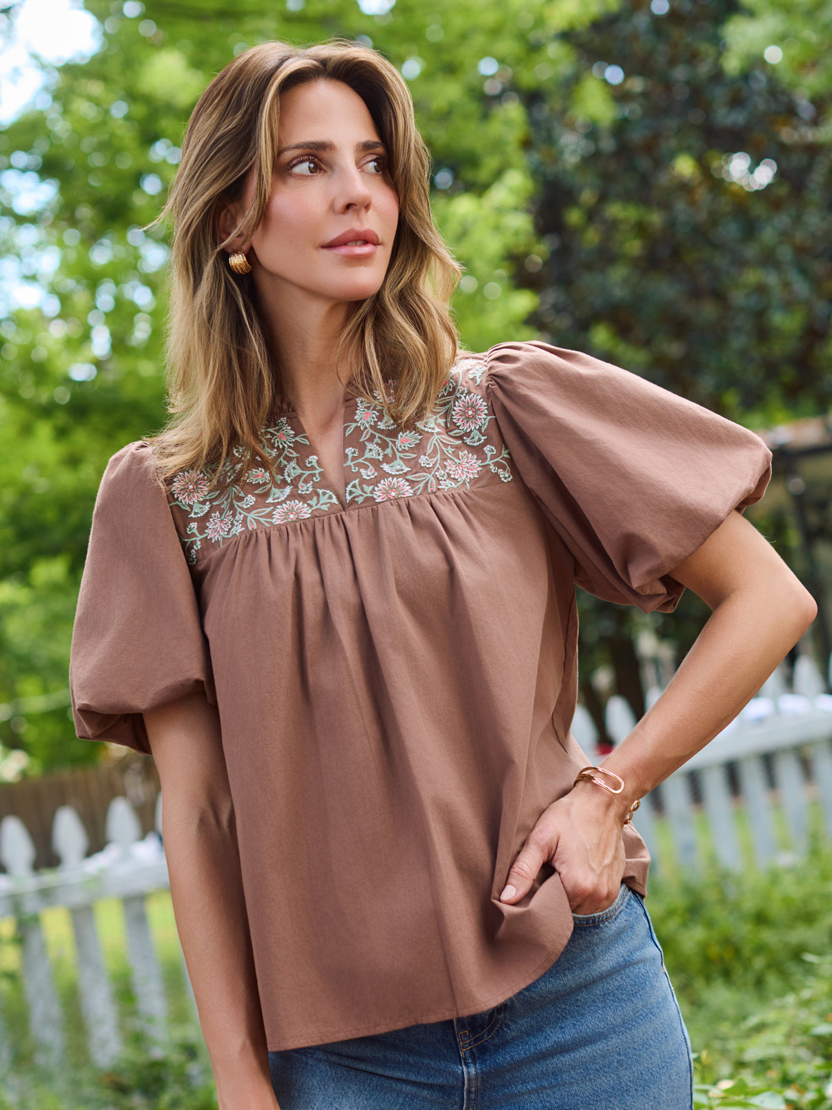 A woman wearing a brown short-sleeve blouse with a floral embroidered yoke and blue jeans, standing outdoors in front of a white picket fence and green trees.