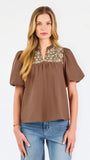 A woman modeling a brown short-sleeve blouse with a floral-patterned yoke and split neckline, paired with blue jeans, against a plain white background.