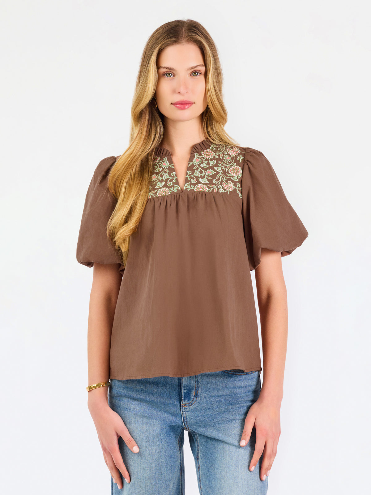 A woman modeling a brown short-sleeve blouse with a floral-patterned yoke and split neckline, paired with blue jeans, against a plain white background.
