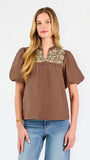 A woman modeling a brown short-sleeve blouse with a floral-patterned yoke and split neckline, paired with blue jeans, against a plain white background.