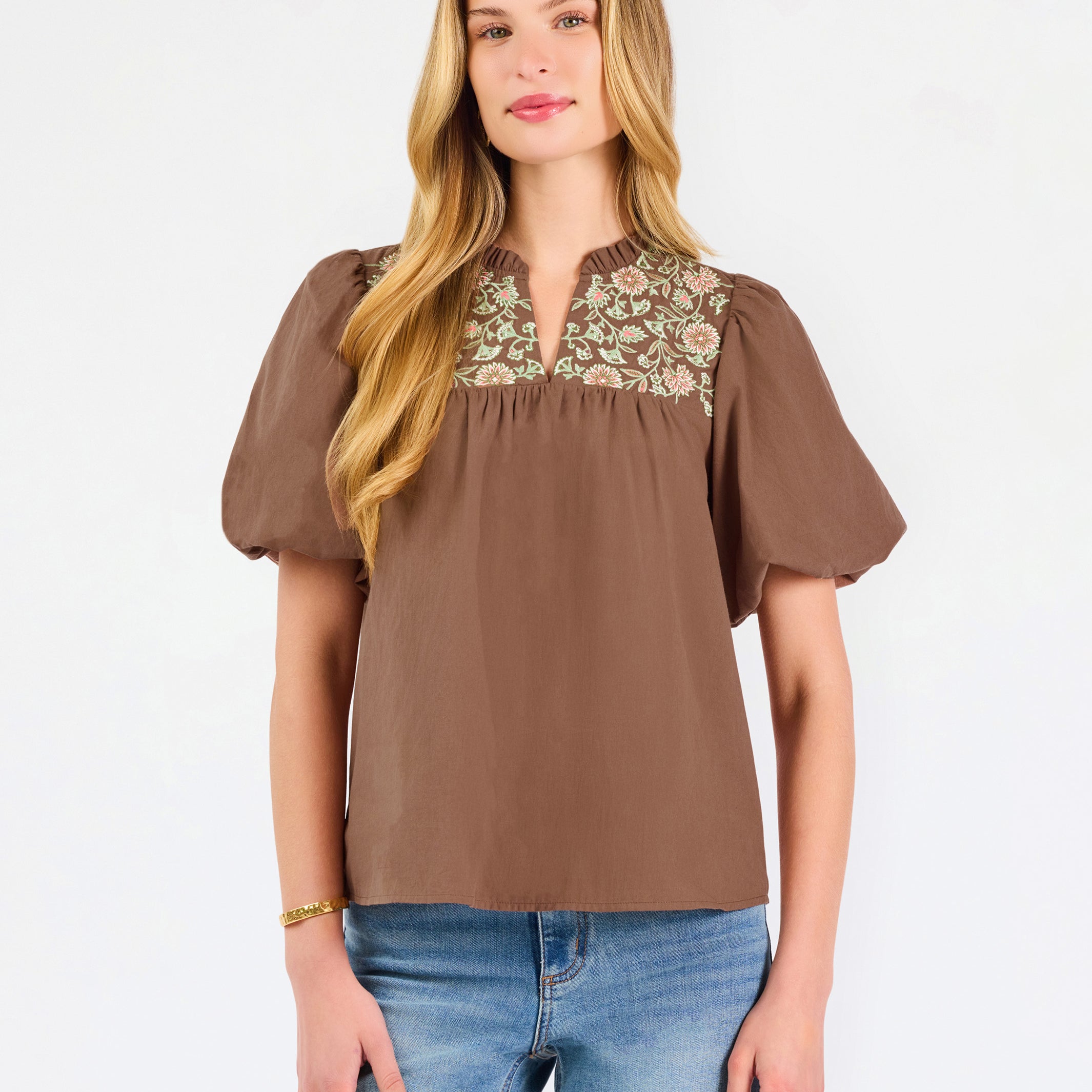 A woman modeling a brown short-sleeve blouse with a floral-patterned yoke and split neckline, paired with blue jeans, against a plain white background.
