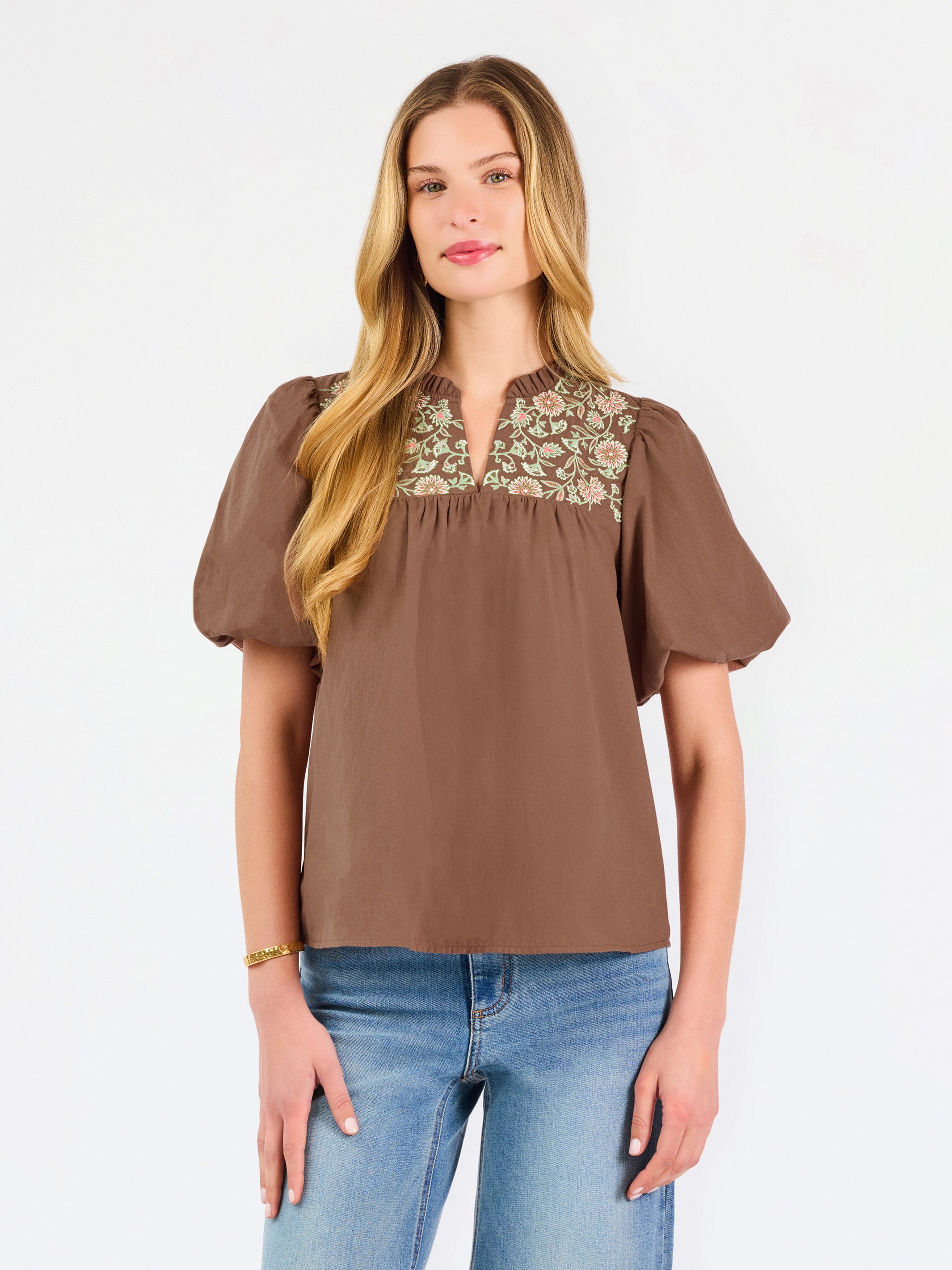 A woman modeling a brown short-sleeve blouse with a floral-patterned yoke and split neckline, paired with blue jeans, against a plain white background.