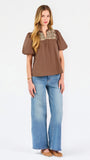 A woman modeling a brown short-sleeve blouse with a floral-patterned yoke and split neckline, paired with blue jeans, against a plain white background.
