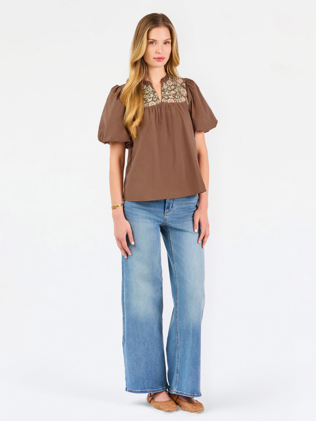 A woman modeling a brown short-sleeve blouse with a floral-patterned yoke and split neckline, paired with blue jeans, against a plain white background.