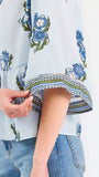 A woman modeling a light blue blouse with blue and green floral embroidery and patterned sleeve cuffs, paired with blue jeans, against a plain white studio background.