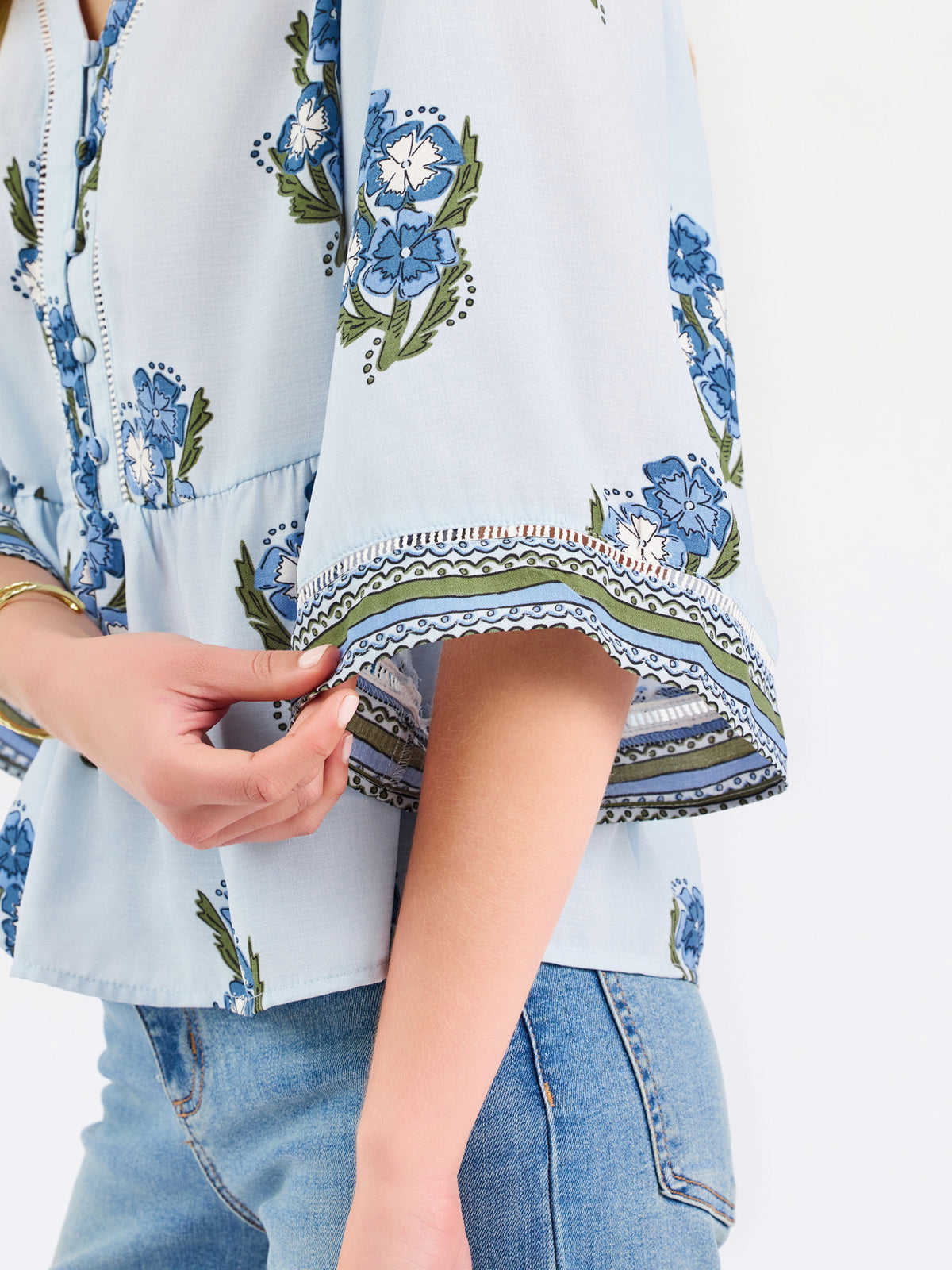 A woman modeling a light blue blouse with blue and green floral embroidery and patterned sleeve cuffs, paired with blue jeans, against a plain white studio background.