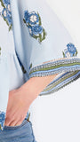 A woman modeling a light blue blouse with blue and green floral embroidery and patterned sleeve cuffs, paired with blue jeans, against a plain white studio background.
