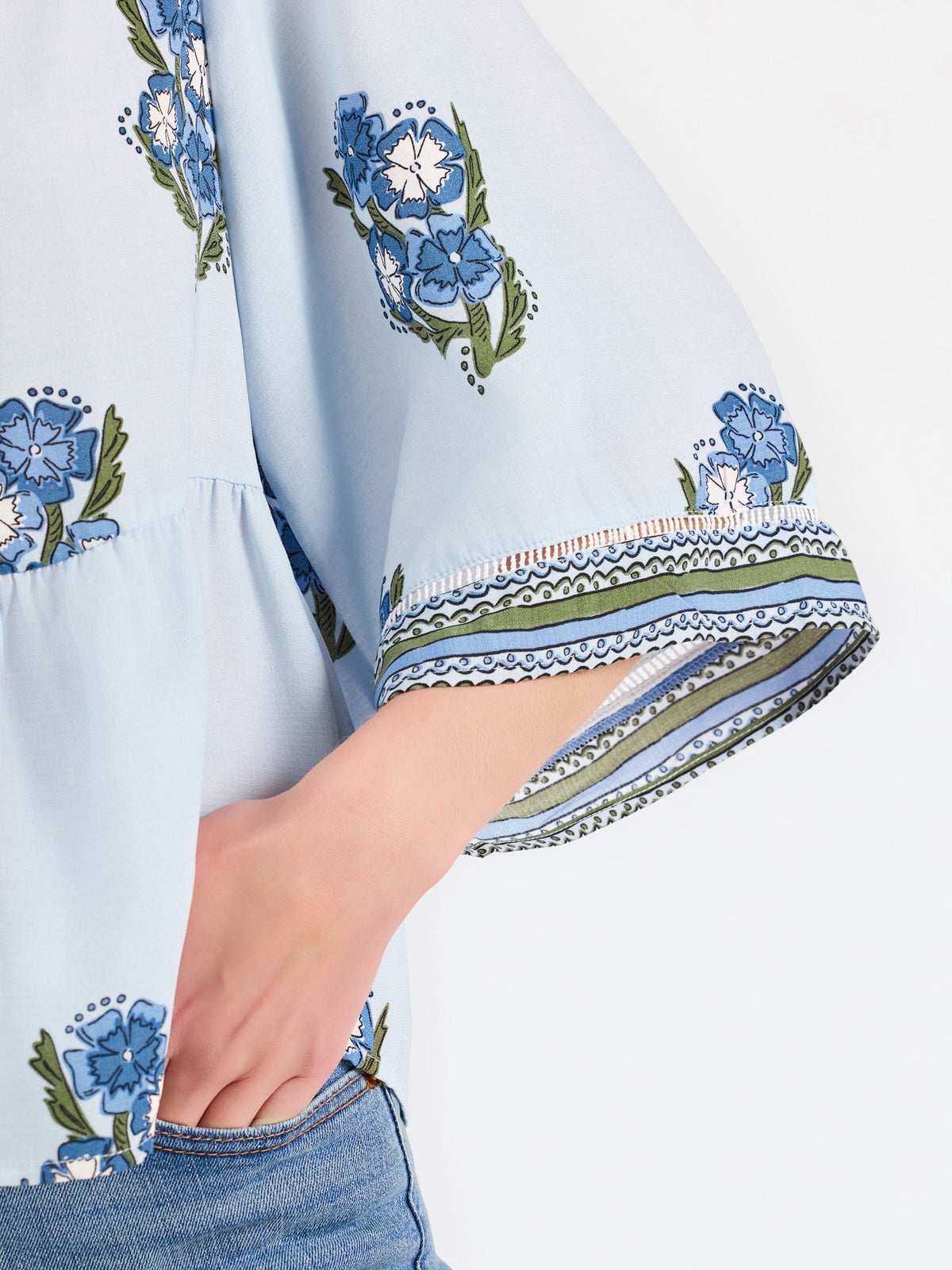 A woman modeling a light blue blouse with blue and green floral embroidery and patterned sleeve cuffs, paired with blue jeans, against a plain white studio background.