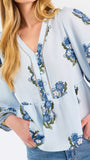 A woman modeling a light blue blouse with blue and green floral embroidery and patterned sleeve cuffs, paired with blue jeans, against a plain white studio background.