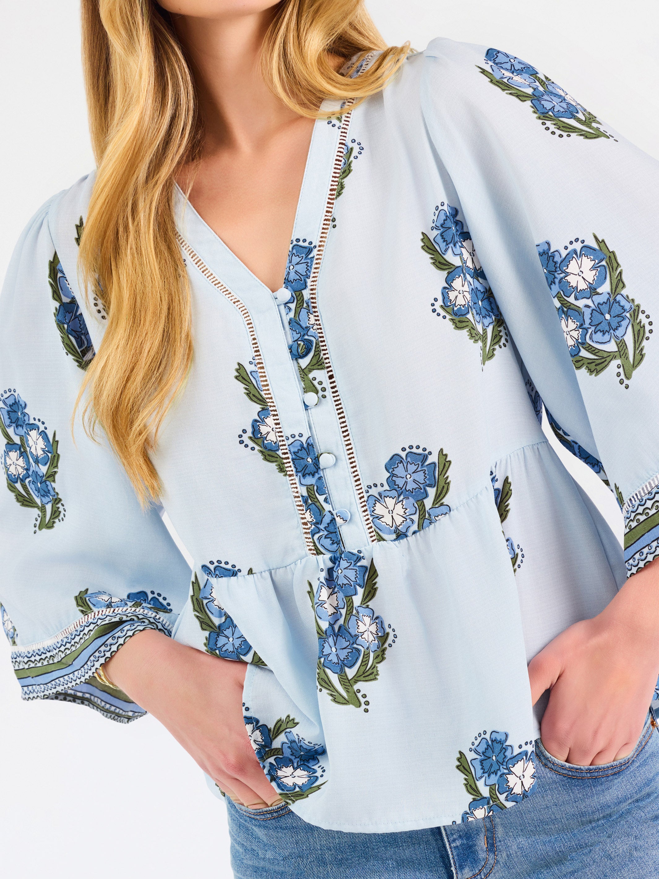 A woman modeling a light blue blouse with blue and green floral embroidery and patterned sleeve cuffs, paired with blue jeans, against a plain white studio background.