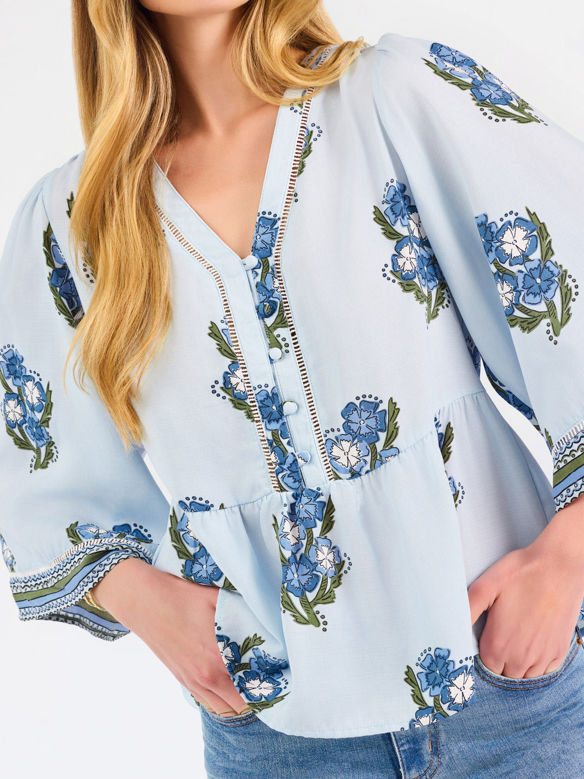 A woman modeling a light blue blouse with blue and green floral embroidery and patterned sleeve cuffs, paired with blue jeans, against a plain white studio background.