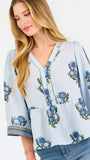 A woman modeling a light blue blouse with blue and green floral embroidery and patterned sleeve cuffs, paired with blue jeans, against a plain white studio background.