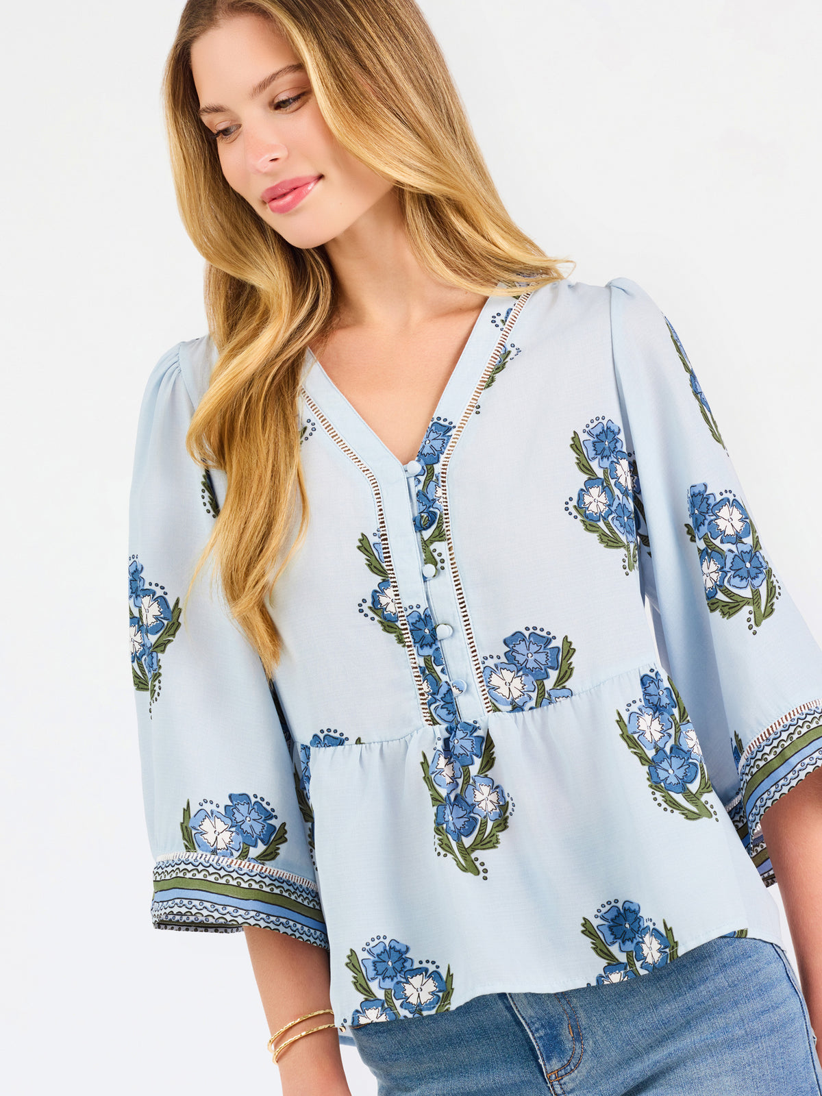 A woman modeling a light blue blouse with blue and green floral embroidery and patterned sleeve cuffs, paired with blue jeans, against a plain white studio background.