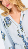 A woman modeling a light blue blouse with blue and green floral embroidery and patterned sleeve cuffs, paired with blue jeans, against a plain white studio background.