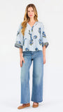 A woman modeling a light blue blouse with blue and green floral embroidery and patterned sleeve cuffs, paired with blue jeans, against a plain white studio background.