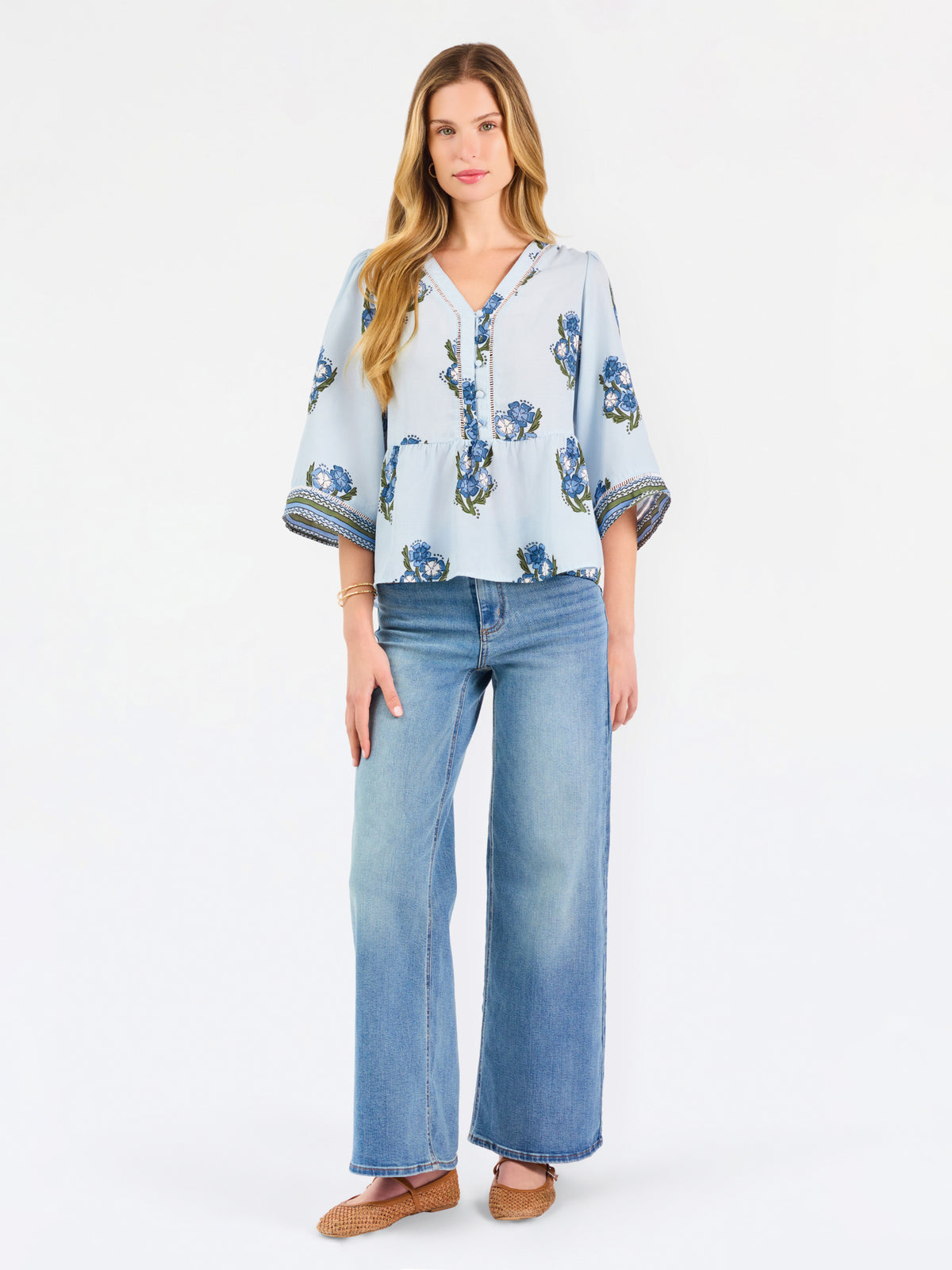 A woman modeling a light blue blouse with blue and green floral embroidery and patterned sleeve cuffs, paired with blue jeans, against a plain white studio background.