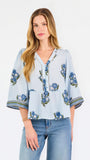 A woman modeling a light blue blouse with blue and green floral embroidery and patterned sleeve cuffs, paired with blue jeans, against a plain white studio background.