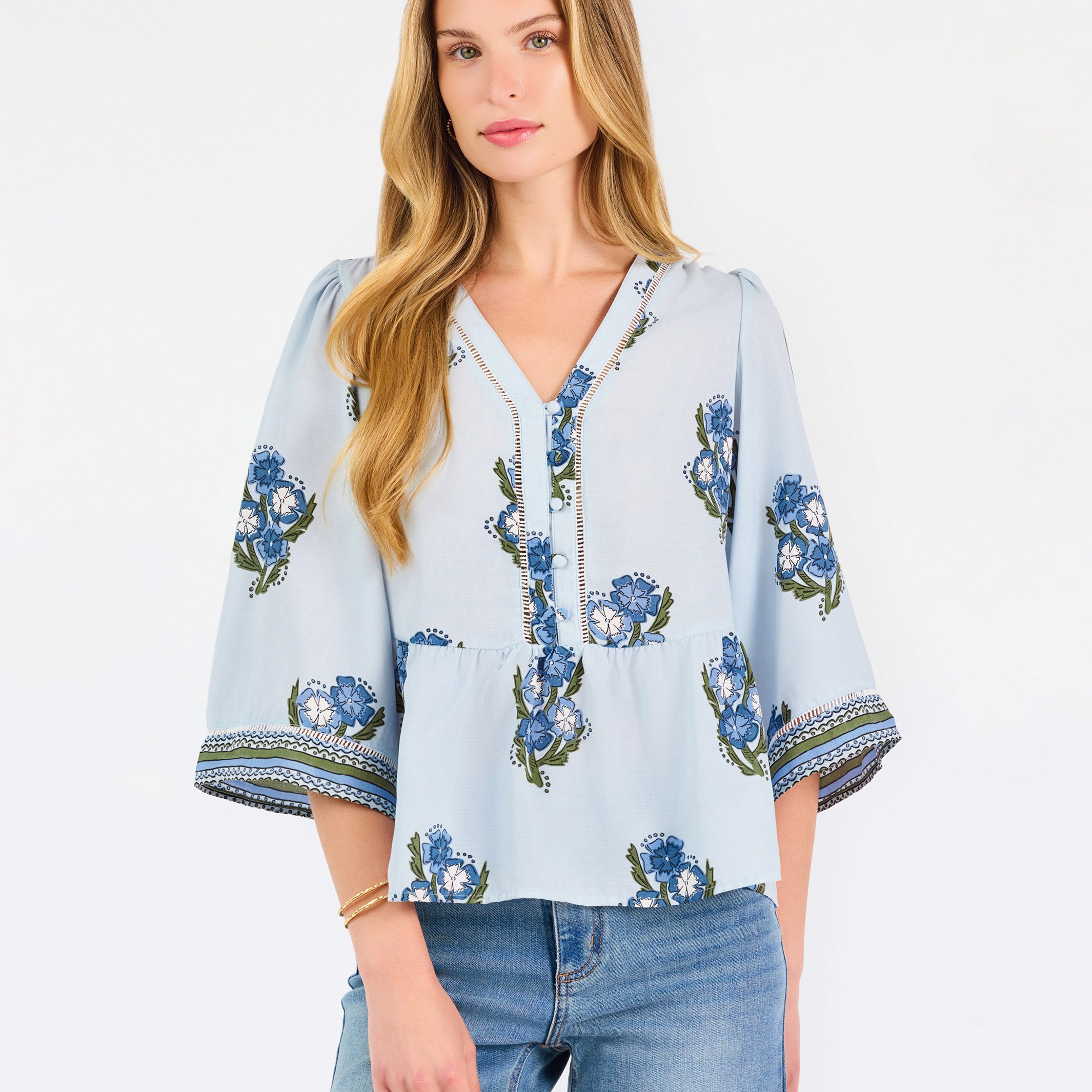 A woman modeling a light blue blouse with blue and green floral embroidery and patterned sleeve cuffs, paired with blue jeans, against a plain white studio background.