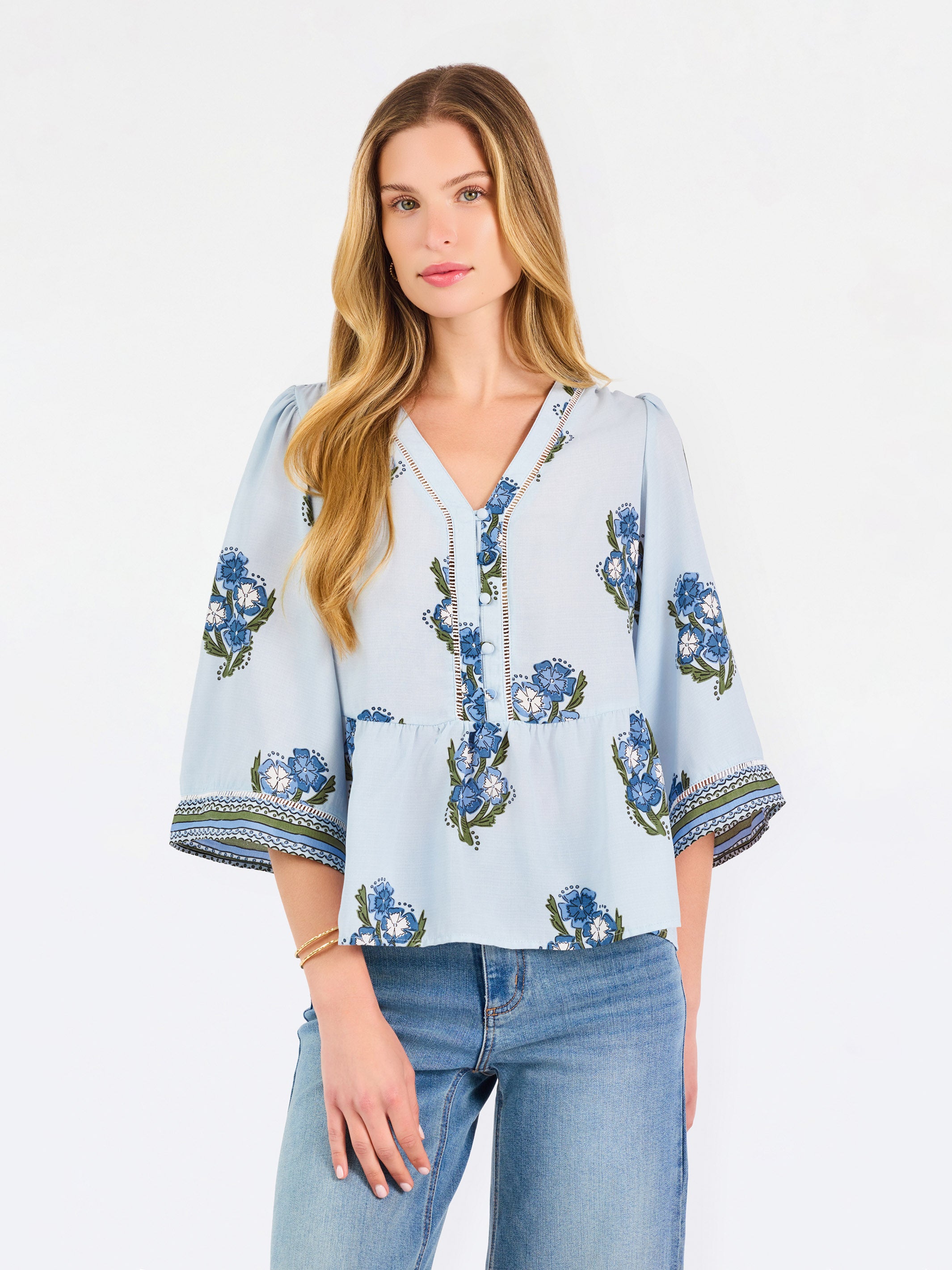 A woman modeling a light blue blouse with blue and green floral embroidery and patterned sleeve cuffs, paired with blue jeans, against a plain white studio background.