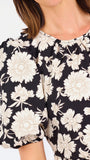 A woman modeling a black puff-sleeve blouse with a large white floral pattern, paired with blue jeans, against a plain white studio background.