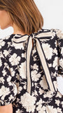 Close-up back view of a woman wearing a black top with a large cream floral print and a tied bow at the neckline.