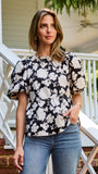 A woman wearing a black and white floral puff-sleeve  top, standing on outdoor brick steps in front of a white porch railing.