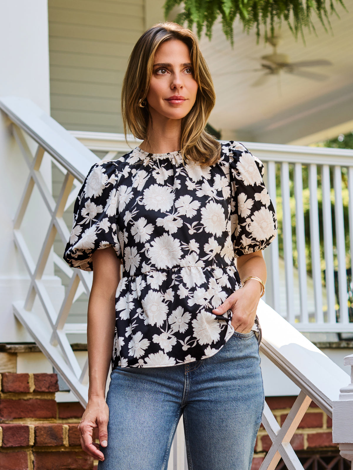 A woman wearing a black and white floral puff-sleeve  top, standing on outdoor brick steps in front of a white porch railing.