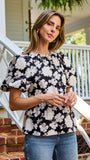 A woman wearing a black and white floral puff-sleeve  top, standing on outdoor brick steps in front of a white porch railing.