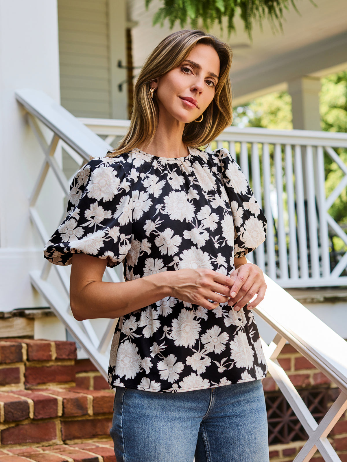 A woman wearing a black and white floral puff-sleeve  top, standing on outdoor brick steps in front of a white porch railing.