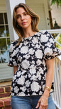 A woman wearing a black and white floral puff-sleeve  top, standing on outdoor brick steps in front of a white porch railing.