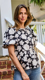 A woman wearing a black puff-sleeve blouse with a white floral pattern, paired with blue jeans, standing on brick steps in front of a white house porch.