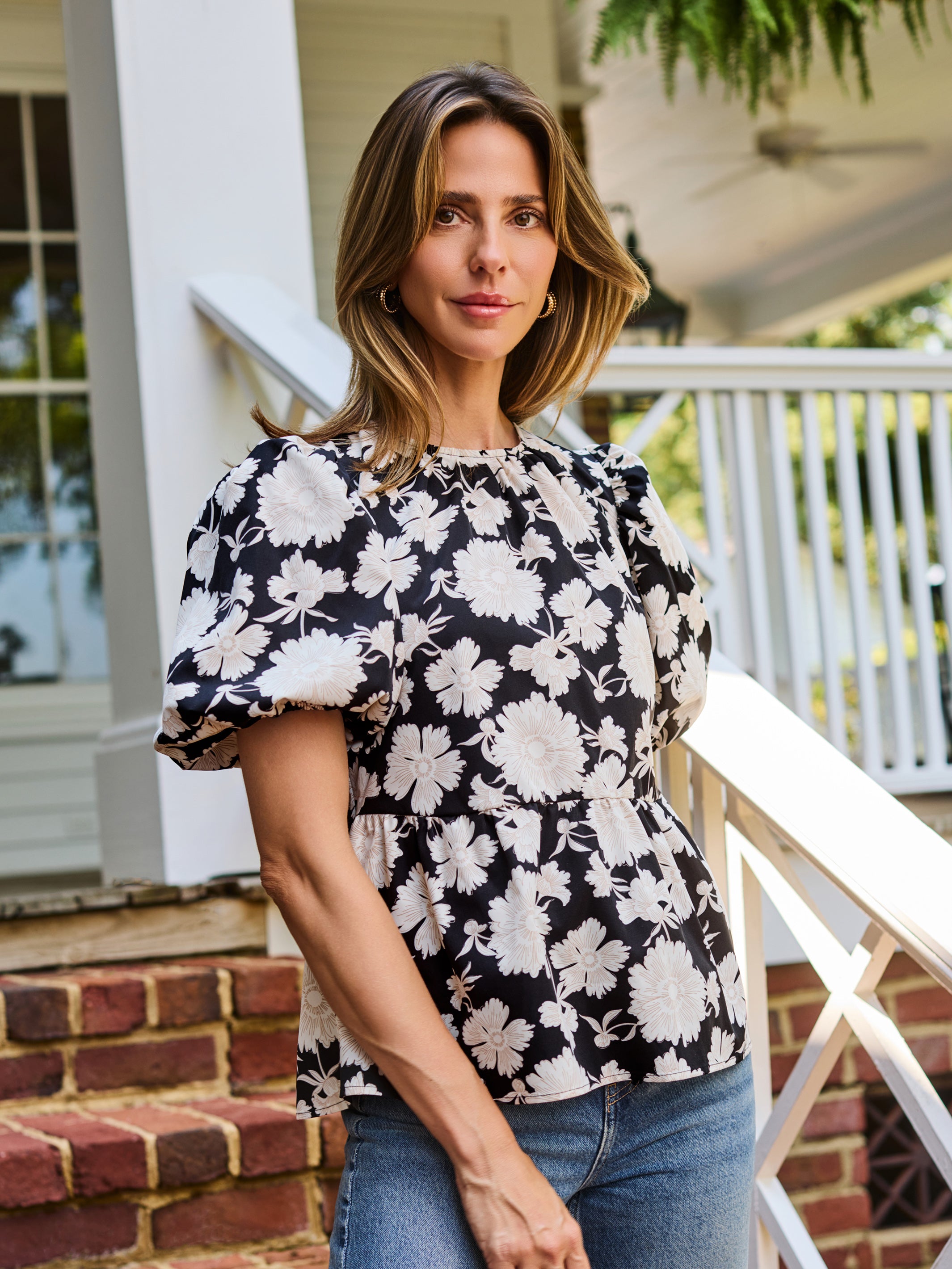 A woman wearing a black puff-sleeve blouse with a white floral pattern, paired with blue jeans, standing on brick steps in front of a white house porch.