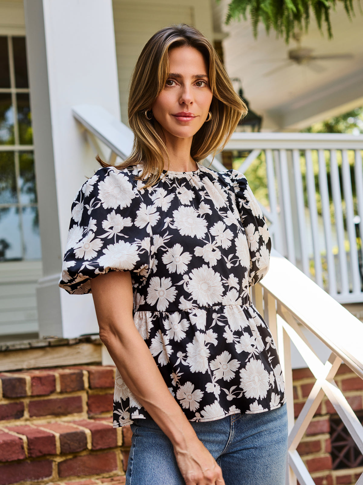 A woman wearing a black puff-sleeve blouse with a white floral pattern, paired with blue jeans, standing on brick steps in front of a white house porch.