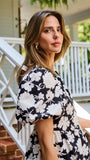 A woman wearing a black and white floral puff-sleeve  top, standing on outdoor brick steps in front of a white porch railing.